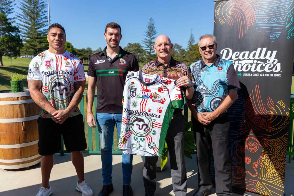 Four men smile in front of a Deadly Choices sign. A man in the middle is holding up his Wynnum Manly Seagulls x Deadly Choices jersey.