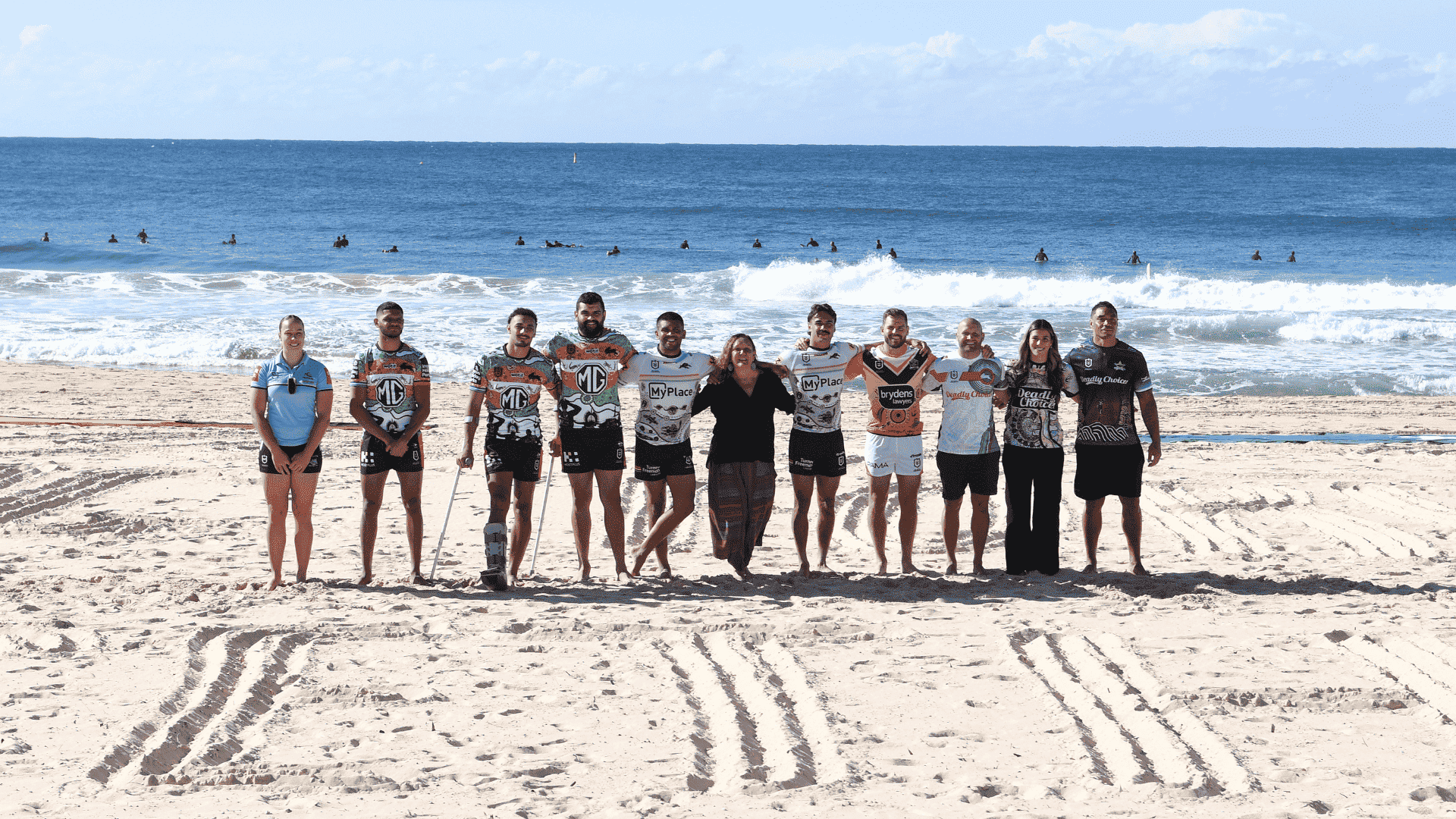 A group of Aboriginal and Torres Strait Islander people stand on the beach, posing with their arms around each other. They are proudly wearing their Deadly Choices jerseys.