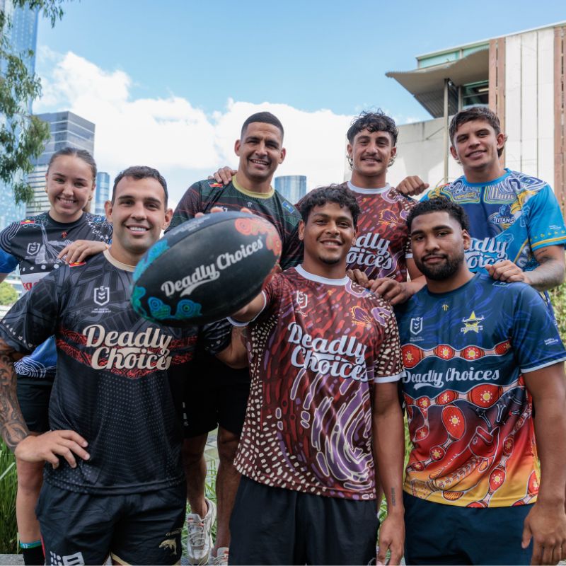 Seven NRL players pose together wearing Deadly Choices jerseys. A male player in the middle holds out a Deadly Choices-branded football.