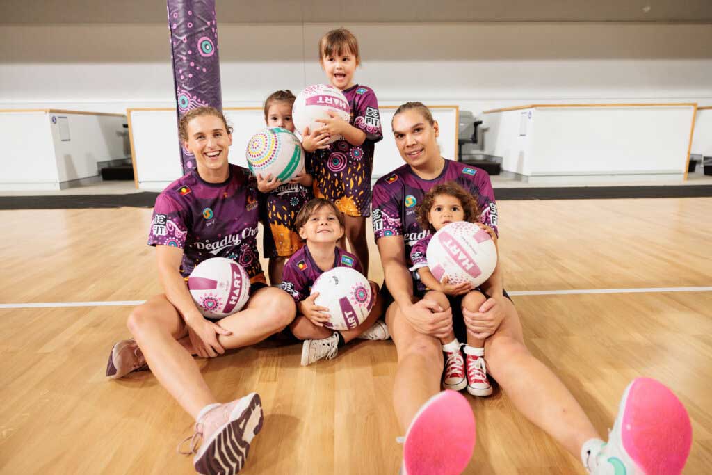 Queensland Firebirds players Gabi Simpson and Donnell Wallam sit on a netball court with young children, all wearing Deadly Choices shirts and holding netballs.