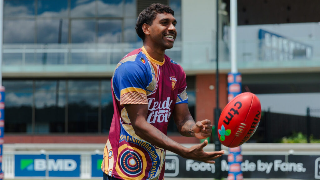 Keidean Coleman wearing a Brisbane Lions Deadly Choices Health Check shirt at the training facility, handballing to a teammate in front of the home goals.