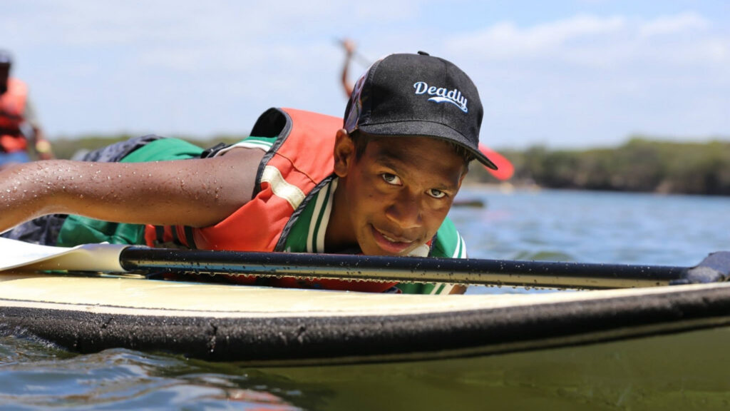 Keidean Coleman wearing a Deadly Choices hat while paddling on a stand up paddle board on the water.