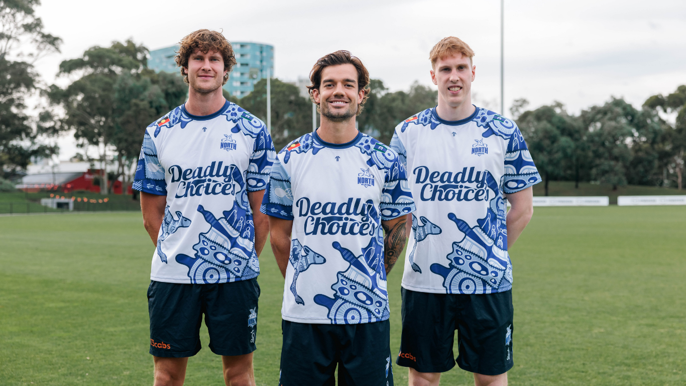 3 sports players standing in a V formation wearing blue and white Deadly Choices t-shirts on an AFL field.